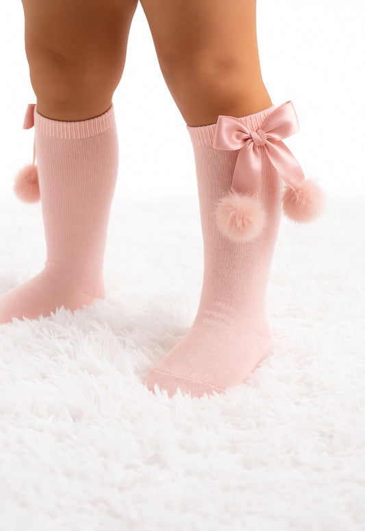 Pink socks with bows and pom-poms worn by a child on a white fluffy surface