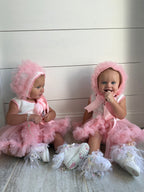 Two babies in pink tutu dresses sitting on a light-colored floor.