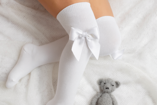 White knee-high socks with bows worn by a person on a soft surface with a teddy bear toy.