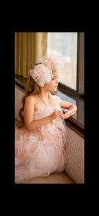 A young girl wearing a light pink ruffled tulle dress and a matching pink feathered headpiece sits on a cushioned window seat, looking out toward a bright window.