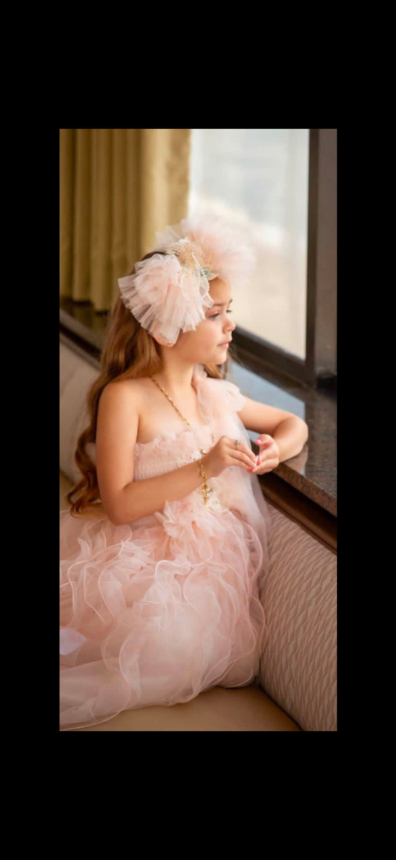 A young girl wearing a light pink ruffled tulle dress and a matching pink feathered headpiece sits on a cushioned window seat, looking out toward a bright window.