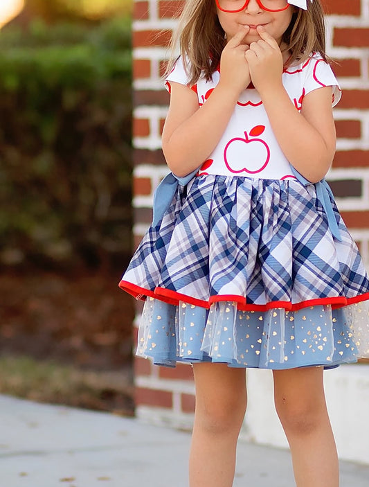 Child wearing a dress with an apple design and plaid skirt in front of a brick wall.