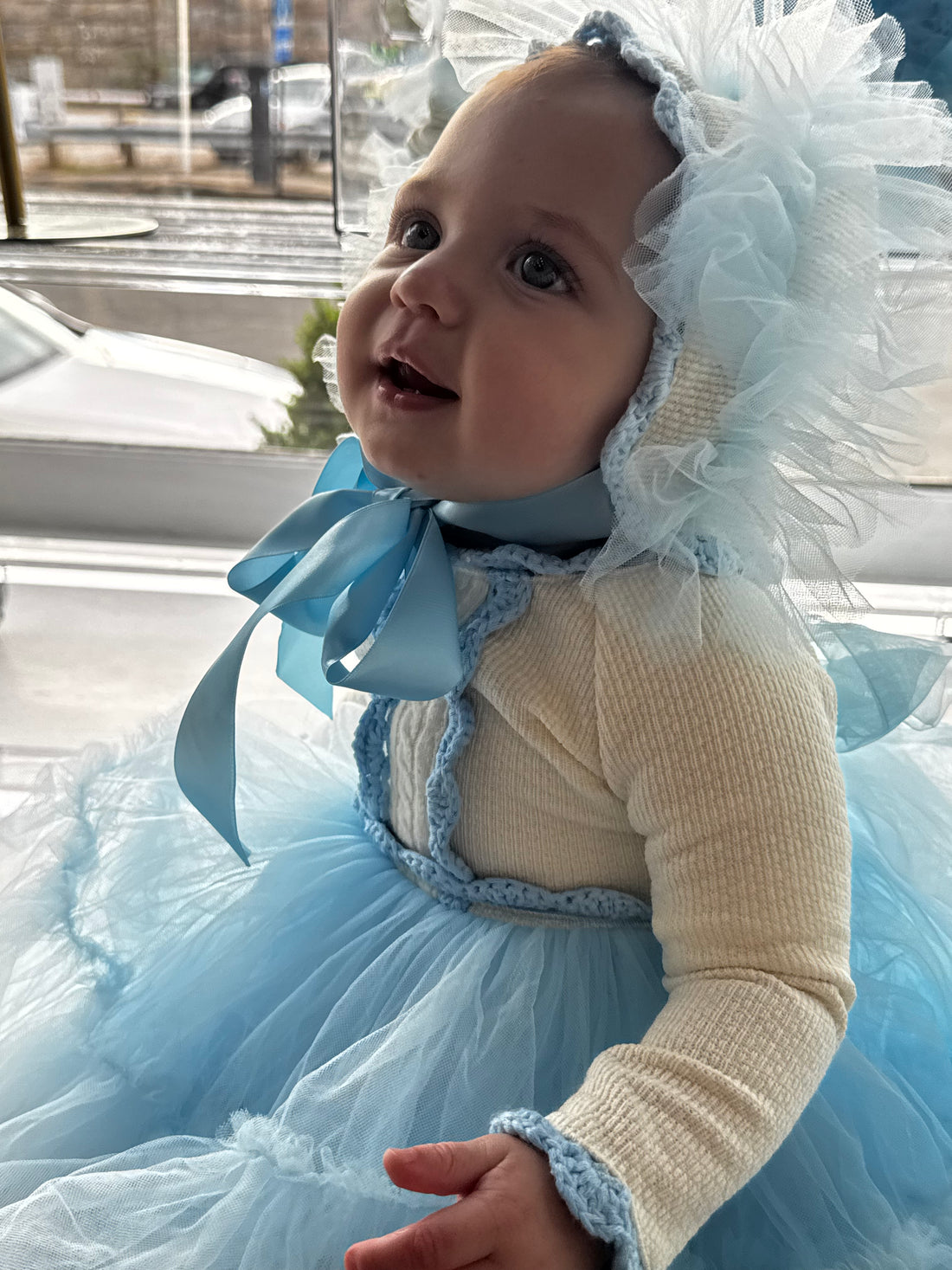 Baby in a blue and white dress with a fluffy headband sitting by a window.