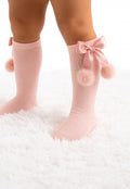 Pink socks with bows and pom-poms worn by a child on a white fluffy surface