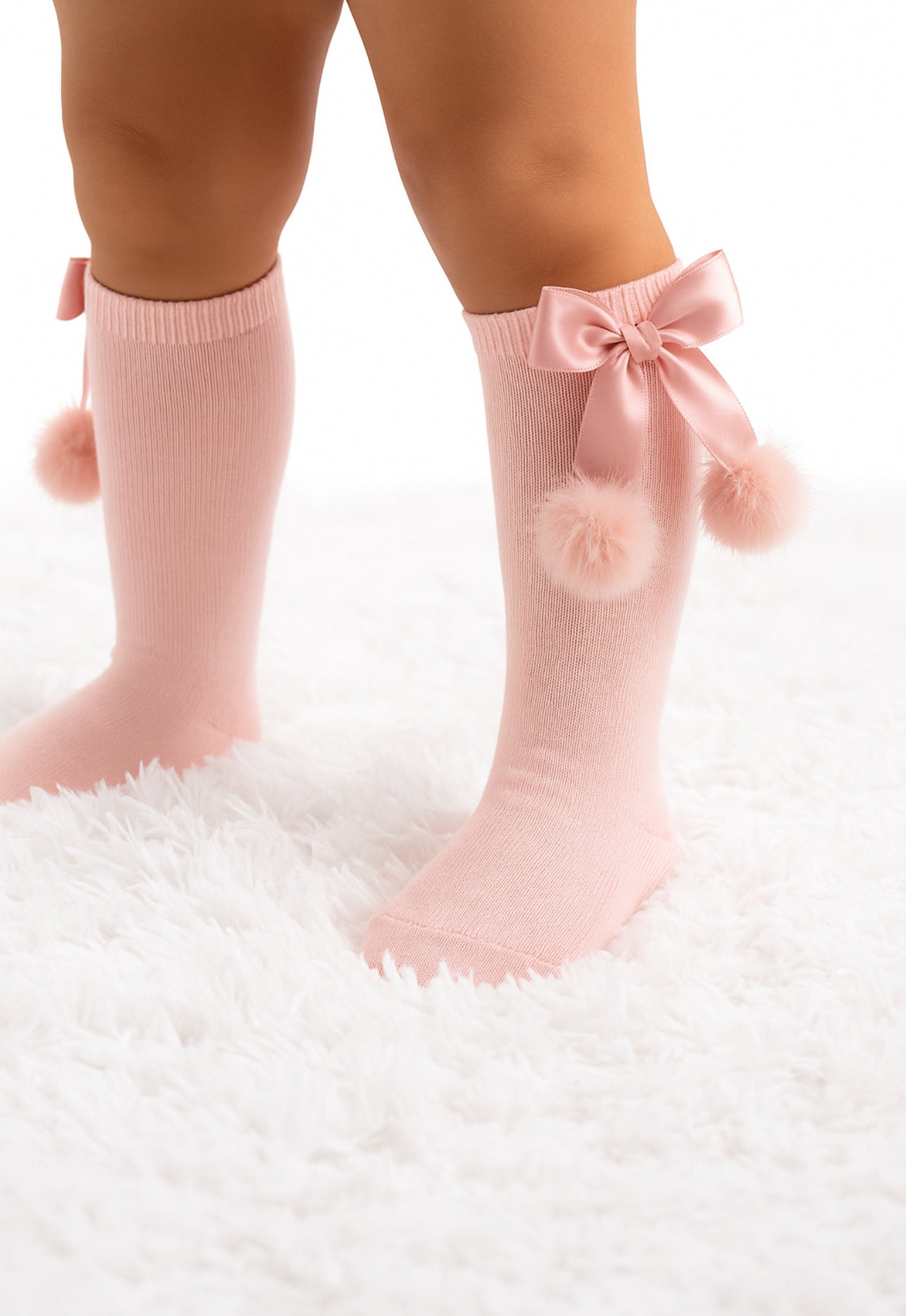 Pink socks with bows and pom-poms worn by a child on a white fluffy surface